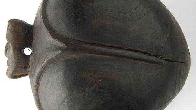 Tahonga: Objeto de madera que se utiliza como adorno, de forma ovoidal, presenta una decoración en alto relieve que la divide en cuatro zonas y en su parte superior posee un apéndice con rostro antropomorfo cruzado por una perforación. Color café oscuro.